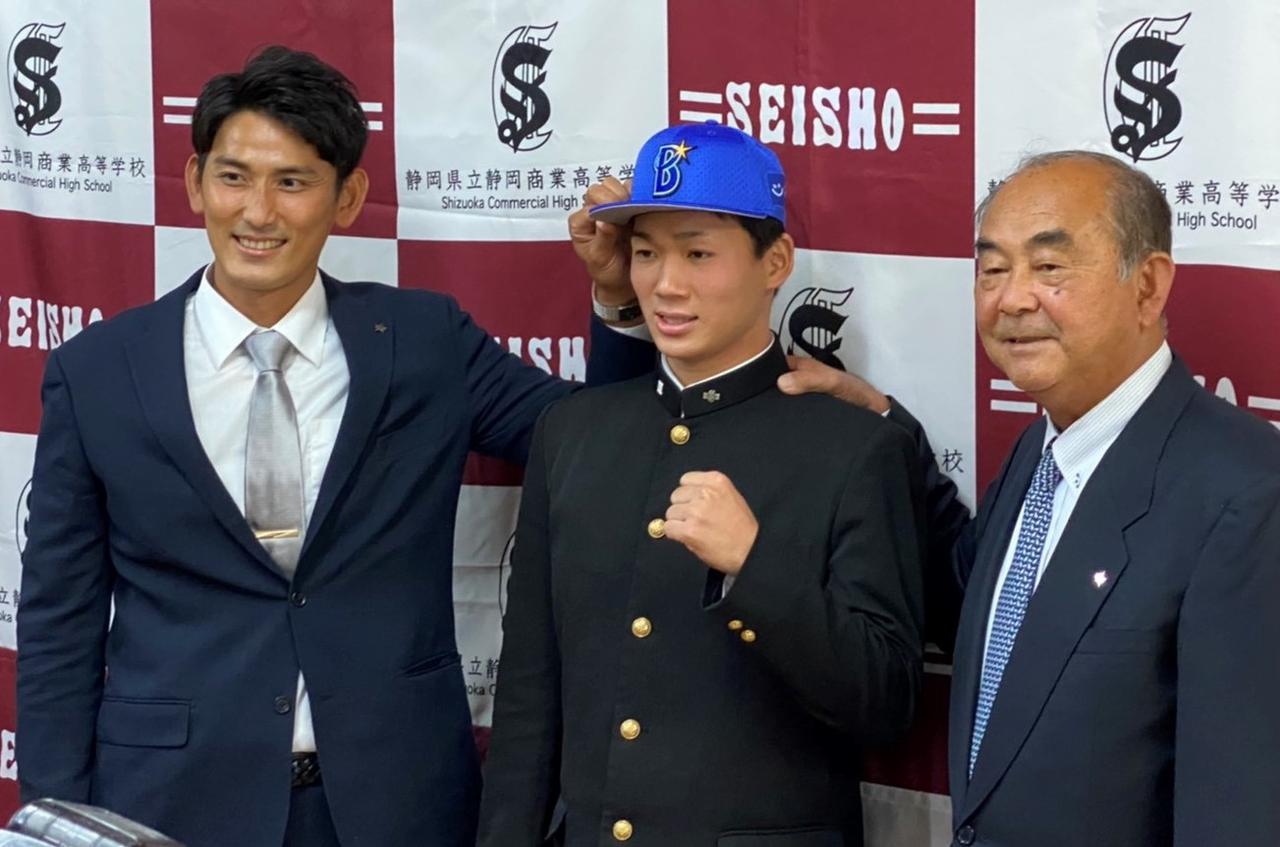 画像: ドラフト指名あいさつを受け、中山大志担当スカウト(左)にDeNaの帽子を被せてもらう高田琢登投手。右は吉田孝司顧問兼代表補佐